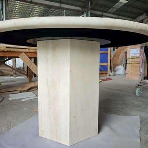 May include: A round dining table featuring a thick, light beige stone top with a smooth, rounded edge. The table is supported by a central, hexagonal pedestal base made of the same light beige stone, accented by a black circular inlay.