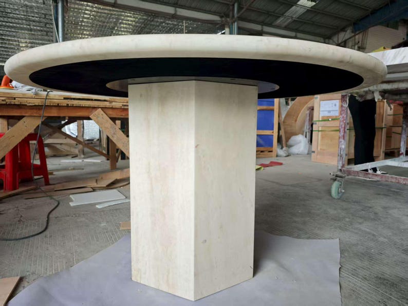 May include: A round dining table featuring a thick, light beige stone top with a smooth, rounded edge. The table is supported by a central, hexagonal pedestal base made of the same light beige stone, accented by a black circular inlay.
