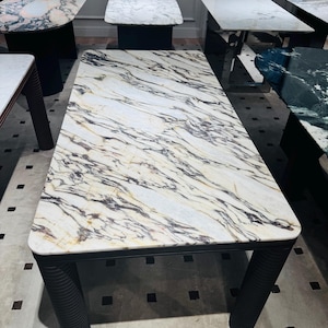 Può includere: Tavolino rettangolare con piano in marmo bianco venato di marrone e nero. La base e le gambe sono nere. Le gambe sono cilindriche e scanalate. Altri tavoli in marmo sono visibili sullo sfondo.