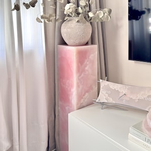 Może przedstawiać: Różowy, prostokątny cokół z teksturowanym, kulistym wazonem z suszonymi liśćmi. Zegar dekoracyjny z chmurą i cyframi 3, 6, 9 i 12 znajduje się na białej szafce. Pokój ma zasłony w neutralnych kolorach.