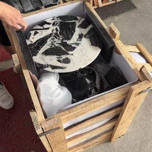 Puede incluir: Una caja de madera que contiene productos de m&aacute;rmol. Se ve una encimera redonda de m&aacute;rmol con vetas negras, junto con otras piezas de m&aacute;rmol. La caja es de madera clara y est&aacute; parcialmente abierta.