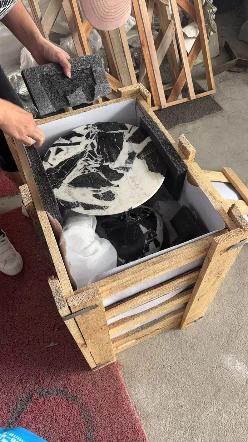 Puede incluir: Una caja de madera que contiene productos de m&aacute;rmol. Se ve una encimera redonda de m&aacute;rmol con vetas negras, junto con otras piezas de m&aacute;rmol. La caja es de madera clara y est&aacute; parcialmente abierta.
