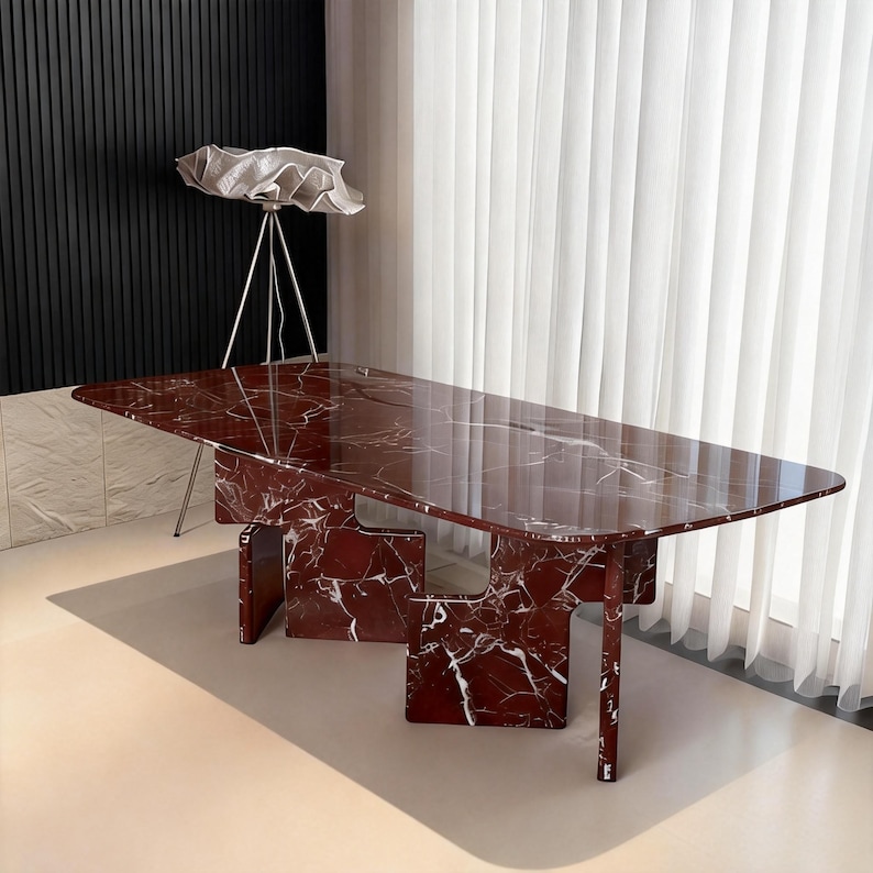 May include: A rectangular, red marble table with white veining. The table has a unique base design with geometric shapes. The table is a statement piece for a modern living space. A white lamp with a unique shade is in the background.