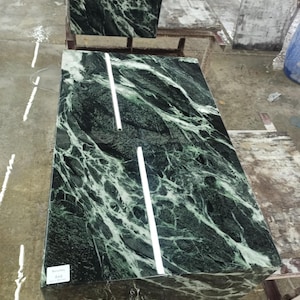Puede incluir: Losas de m&aacute;rmol verde con vetas blancas. Las losas rectangulares tienen una superficie pulida. Dos l&iacute;neas blancas, rectas, finas y verticales est&aacute;n en la losa superior. Las losas est&aacute;n sobre una estructura de madera. Una etiqueta con texto est&aacute; en la losa inferior.