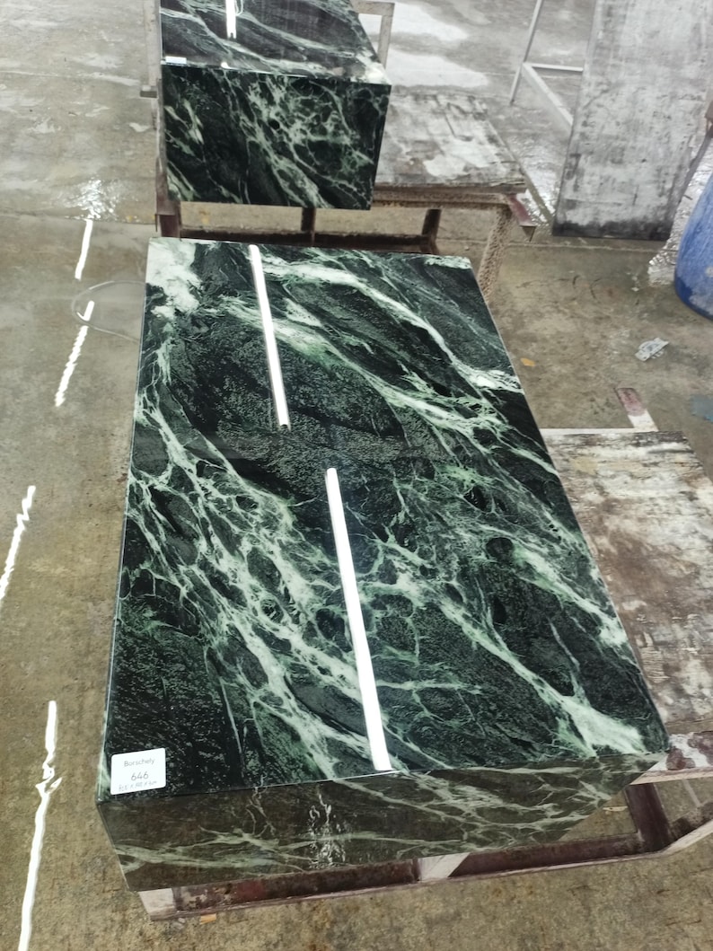 Puede incluir: Losas de m&aacute;rmol verde con vetas blancas. Las losas rectangulares tienen una superficie pulida. Dos l&iacute;neas blancas, rectas, finas y verticales est&aacute;n en la losa superior. Las losas est&aacute;n sobre una estructura de madera. Una etiqueta con texto est&aacute; en la losa inferior.