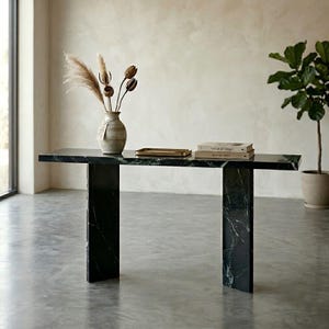 May include: A rectangular black marble console table with a polished surface and two thick vertical slab legs. On the table sits a beige ceramic vase containing dried pampas grass and seed pods, a small gold tray, and two stacked hardcover books.