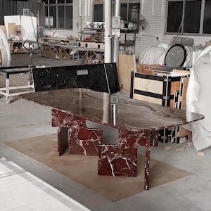 May include: A large, rectangular dining table with a burgundy marble top and matching geometric legs. The table is set on a light brown surface in a warehouse setting. The marble has white veining.