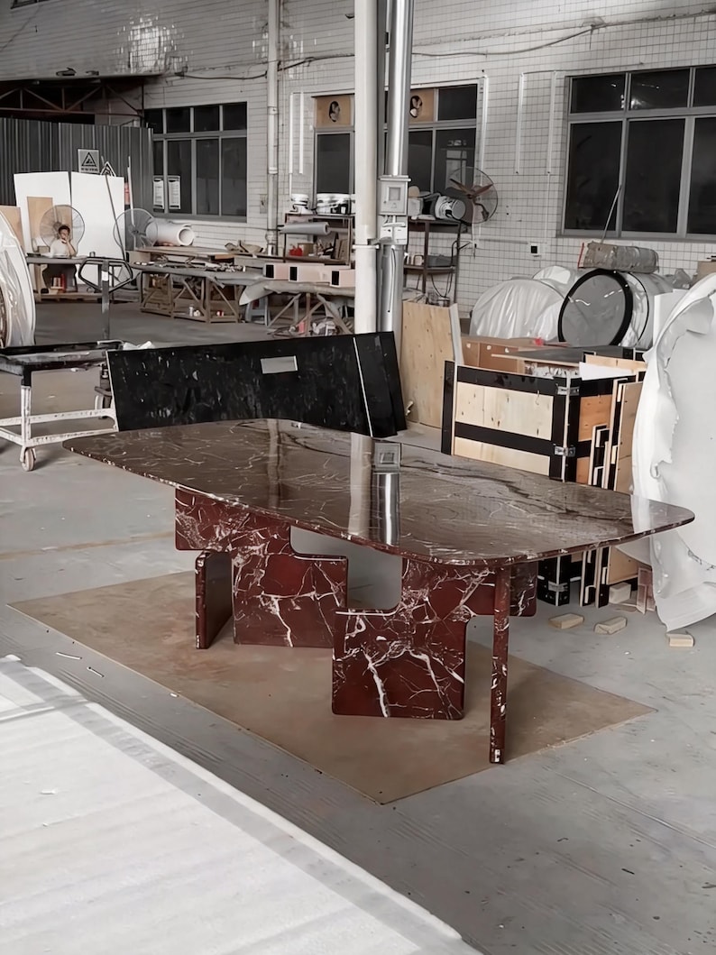 May include: A large, rectangular dining table with a burgundy marble top and matching geometric legs. The table is set on a light brown surface in a warehouse setting. The marble has white veining.