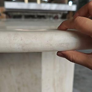 May include: A light beige marble table with a smooth, rounded edge and a polished surface. The stone features subtle, natural veining and a consistent, creamy color palette throughout the structure, showcasing a solid and sturdy construction.