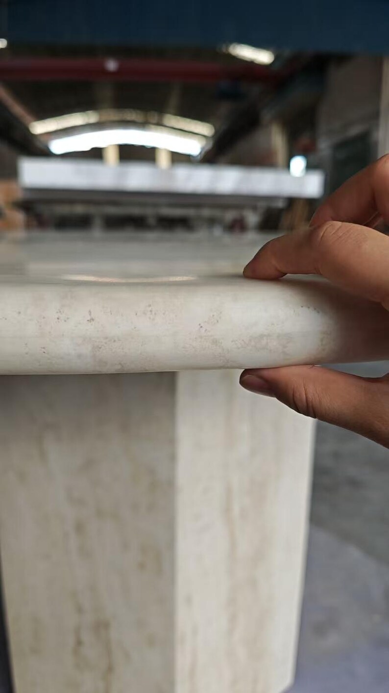 May include: A light beige marble table with a smooth, rounded edge and a polished surface. The stone features subtle, natural veining and a consistent, creamy color palette throughout the structure, showcasing a solid and sturdy construction.