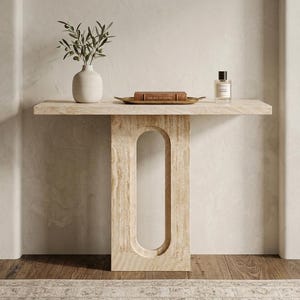 Op de afbeelding: Een lichtgekleurde, rechthoekige console met een langwerpige ovale uitsparing in het midden. De tafel is versierd met een vaas met groen, een boek op een dienblad en een parfumflesje. De tafel is gemaakt van lichtgekleurde steen of hout.