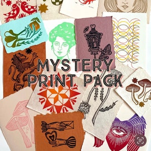 May include: A collection of 12 unique, hand-printed art prints featuring various designs, including a lion, a star, a lantern, a mushroom, and a hand holding a heart. The prints are on different colored papers and have a vintage, handmade aesthetic. The text "MYSTERY PRINT PACK" is displayed in the center of the image.
