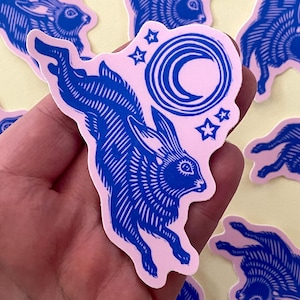 May include: A collection of stickers featuring a blue rabbit leaping towards a crescent moon and stars on a pink background. The stickers have a linocut style and are arranged on a light yellow surface.