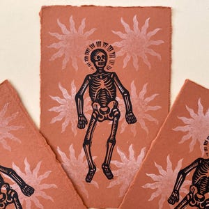 May include: Three handmade art prints. Each features a black skeleton figure with a halo crown, set against a terracotta background with white sunburst designs. The prints have a textured, deckled edge, giving them a rustic feel.