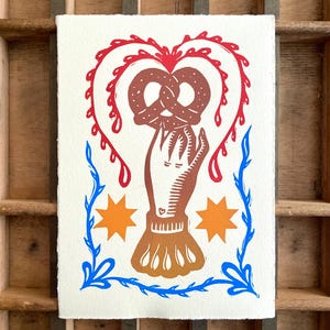 May include: A print featuring a hand holding a pretzel, surrounded by a red heart-shaped design and blue floral accents. Two orange starbursts are also included in the design. The artwork is on a white background.