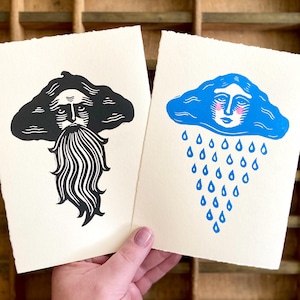 May include: Two art prints held by a hand. One print features a black bearded figure, the other a blue cloud with a face and falling raindrops. Both prints are on off-white paper.