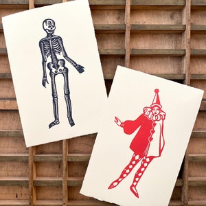 May include: Two vintage woodblock prints on cream paper. The left print is a black skeleton with a skull. The right print is a red jester with a white ruff and a red hat.
