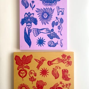 May include: Two wooden panels with linocut prints. The top panel is light purple with blue images of a moth, flowers, and a sun. The bottom panel is gold with red images of a hummingbird, a rabbit, and a beet.