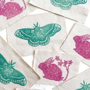 May include: A collection of fabric patches featuring linocut prints of moths and rabbits in teal and magenta. The designs include detailed illustrations of the insects and animals, some with floral accents. The patches are arranged on a white surface.
