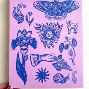 May include: A lilac-colored wooden panel featuring blue linocut illustrations of a moth, flowers, a sun face, a lamb, a hand, a snail, and berries. The artwork has a folk art style.