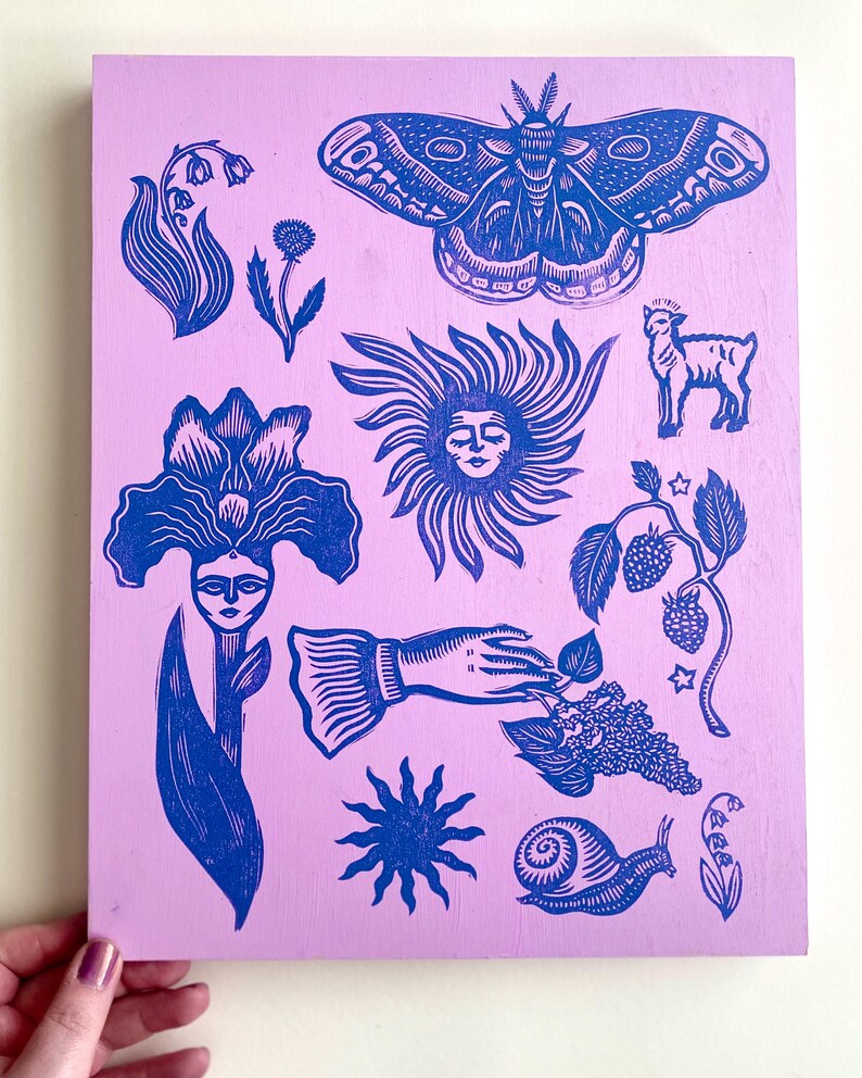 May include: A lilac-colored wooden panel featuring blue linocut illustrations of a moth, flowers, a sun face, a lamb, a hand, a snail, and berries. The artwork has a folk art style.