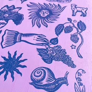 May include: A lilac-colored wooden block with various blue linocut images. The images include a moth, a sun with a face, a lamb, a hand holding a leaf, a snail, flowers, and berries. The overall theme is nature and botanical.