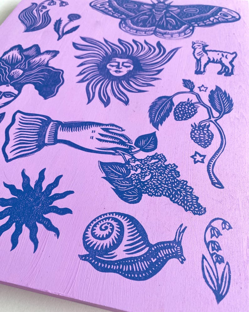 May include: A lilac-colored wooden block with various blue linocut images. The images include a moth, a sun with a face, a lamb, a hand holding a leaf, a snail, flowers, and berries. The overall theme is nature and botanical.