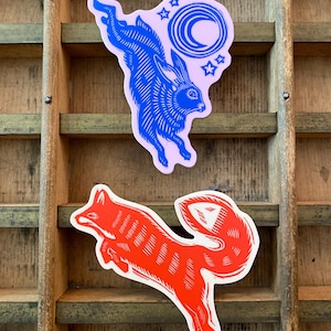 Puede incluir: Dos pegatinas: un conejo azul con una luna y estrellas sobre fondo rosa, y un zorro rojo. Ambas pegatinas tienen un estilo linograbado y est&aacute;n sobre un fondo de madera.