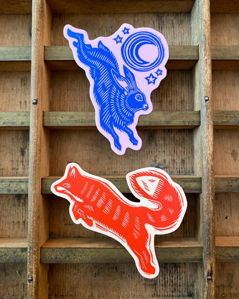 Puede incluir: Dos pegatinas: un conejo azul con una luna y estrellas sobre fondo rosa, y un zorro rojo. Ambas pegatinas tienen un estilo linograbado y est&aacute;n sobre un fondo de madera.