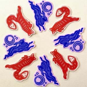Puede incluir: Un conjunto de pegatinas con zorros rojos y conejos azules en poses din&aacute;micas. Cada pegatina tiene un dise&ntilde;o &uacute;nico, con un fondo lila claro que incluye una luna creciente y estrellas. Las pegatinas est&aacute;n dispuestas en un patr&oacute;n circular.