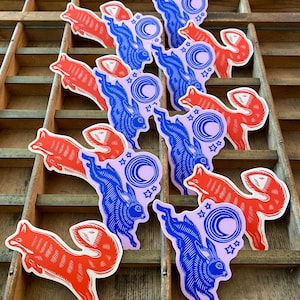 Puede incluir: Colecci&oacute;n de pegatinas con un zorro rojo y un conejo azul con un dise&ntilde;o de luna creciente y estrellas. Las pegatinas est&aacute;n dispuestas sobre una superficie de madera con m&uacute;ltiples compartimentos.