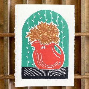 May include: A block print on textured white paper featuring a bright red pitcher filled with orange sunflowers. The pitcher sits on a black surface against a green arched background with white vertical vine patterns and thin white leaf details.
