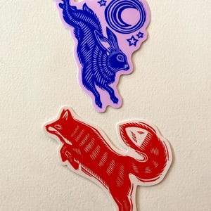 Puede incluir: Dos pegatinas troqueladas. Una presenta un conejo azul saltando sobre un fondo rosa con una luna creciente y estrellas. La otra es un zorro rojo en posici&oacute;n de salto. Ambas pegatinas tienen un borde blanco y est&aacute;n sobre un fondo blanco.