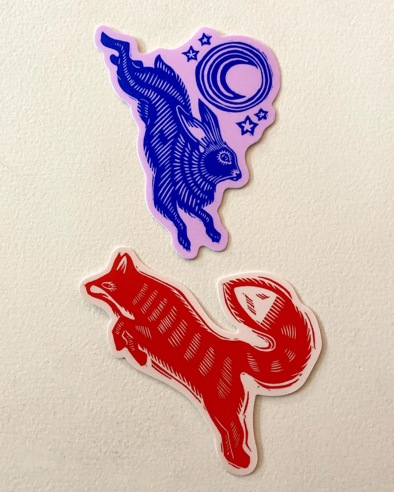 Puede incluir: Dos pegatinas troqueladas. Una presenta un conejo azul saltando sobre un fondo rosa con una luna creciente y estrellas. La otra es un zorro rojo en posici&oacute;n de salto. Ambas pegatinas tienen un borde blanco y est&aacute;n sobre un fondo blanco.