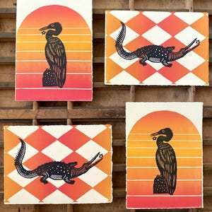 May include: Four art prints with a nature theme. Two prints feature a black bird silhouette against a sunset background. The other two prints show a black alligator on a diamond pattern background in orange and white.