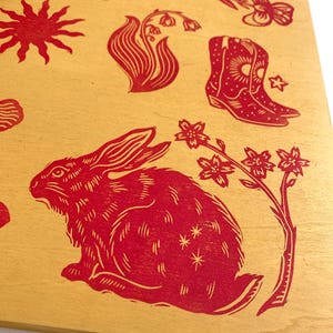 May include: A wooden panel featuring red linocut prints of various designs. The images include a rabbit, flowers, a hand holding flowers, a sun, a hummingbird, a woman's face, and cowboy boots. The background is a light tan color.
