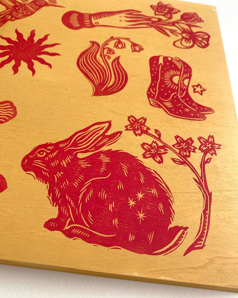 May include: A wooden panel featuring red linocut prints of various designs. The images include a rabbit, flowers, a hand holding flowers, a sun, a hummingbird, a woman's face, and cowboy boots. The background is a light tan color.