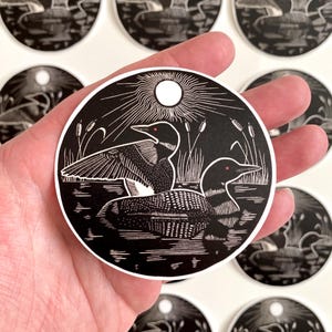 May include: Black and white round sticker featuring two loons on a lake with cattails and a sunburst design. The loons have red eyes. The sticker is held in a hand, with several other stickers visible in the background.