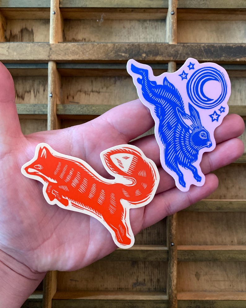 Puede incluir: Dos pegatinas: un zorro rojo y un conejo azul. El zorro es naranja con detalles blancos. El conejo es azul con un fondo rosa, una luna creciente y estrellas. Las pegatinas se sostienen en una mano.