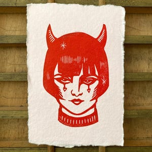 May include: Red linocut print depicting a stylized face with horns, a star, and heart-shaped accents under the eyes. The print is on textured white paper. The artwork is a portrait of a devilish figure.