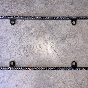 May include: Black license plate frame embellished with sparkling rhinestones. The frame has four mounting holes and the text "Alikajade Designs" is visible. A decorative accessory for vehicles.