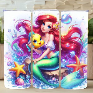Puede incluir: Un vaso blanco con una vibrante ilustración de una sirena con largo cabello rojo, una cola verde y un pez amarillo. El diseño incluye burbujas, estrellas de mar y plantas acuáticas, creando una escena submarina caprichosa.