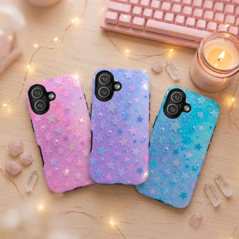 May include: Three phone cases with star patterns in pink, purple, and blue hues. Each case has a black border and a camera cutout. The cases are on a wooden surface with a keyboard, candle, and crystals in the background.