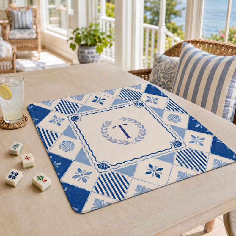 Op de afbeelding: Een blauw-wit patroon placemat met een centraal ontwerp met de letter "T" in een lauwerkrans. De placemat heeft een nautisch thema met schelp- en bloemmotieven. De placemat ligt op een lichtgekleurde houten tafel.