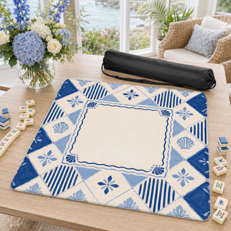Op de afbeelding: Een blauw-wit patroon Mahjong-mat met een cr&egrave;mekleurig midden. De mat heeft een nautisch ontwerp met schelpen en bloemmotieven. Een zwarte draagtas en Mahjong-tegels zijn ook aanwezig. De mat ligt op een lichte houten tafel.