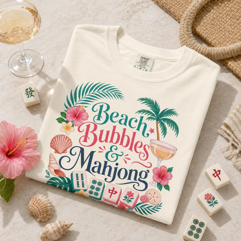May include: Cream-coloured sweatshirt featuring the text "Beach Bubbles & Mahjong" in a vibrant design. The graphic includes palm trees, hibiscus flowers, seashells, and mahjong tiles. A glass of champagne and mahjong tiles are also visible.
