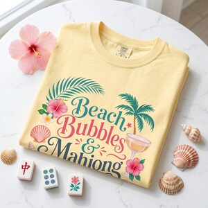 May include: A pale yellow t-shirt with the text "Beach Bubbles & Mahiong" in a tropical design. The shirt features a palm tree, hibiscus flowers, and a cocktail glass. Seashells and Mahjong tiles are arranged around the shirt.