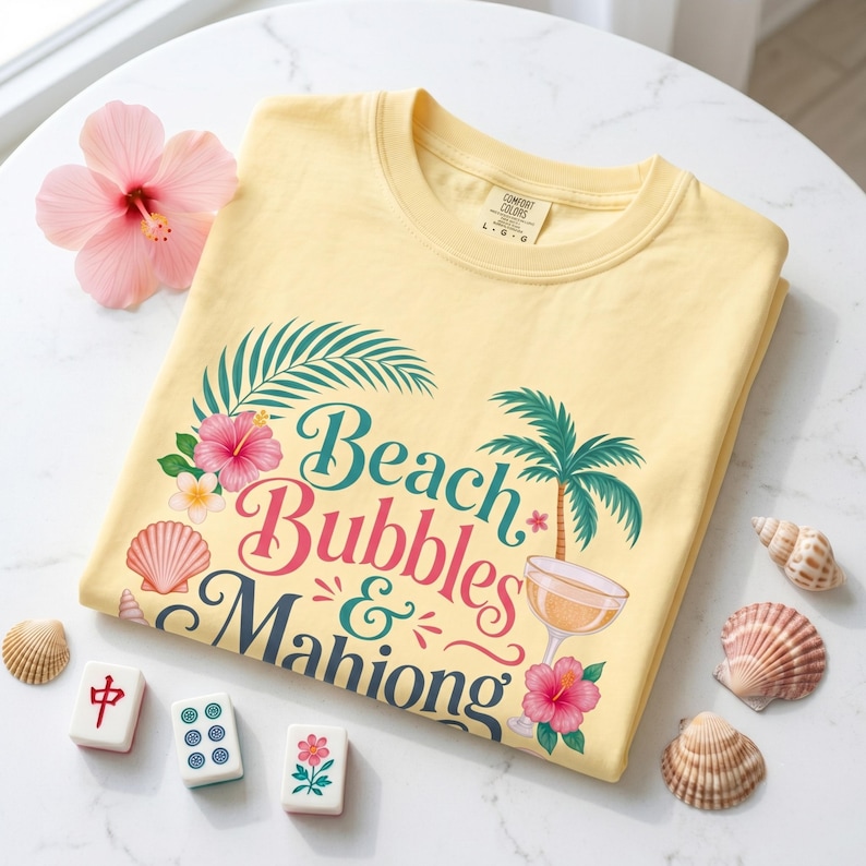 May include: A pale yellow t-shirt with the text "Beach Bubbles & Mahiong" in a tropical design. The shirt features a palm tree, hibiscus flowers, and a cocktail glass. Seashells and Mahjong tiles are arranged around the shirt.