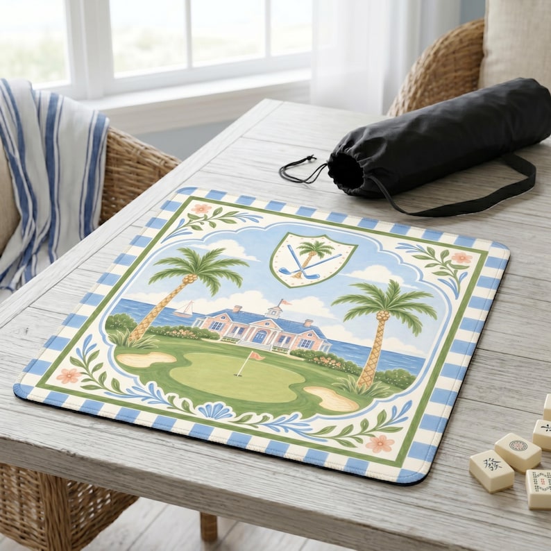 May include: A rectangular golf-themed mousepad featuring a scenic illustration of a golf course with palm trees, a clubhouse, and a blue and white checkered border. A black carrying bag rests on the table.