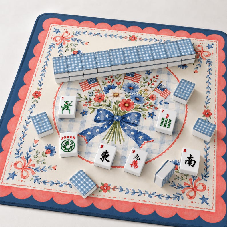 May include: A Mahjong set with blue and white checkered tiles, arranged on a floral patterned mat with a red and blue scalloped border. The mat features a bouquet with American flags.
